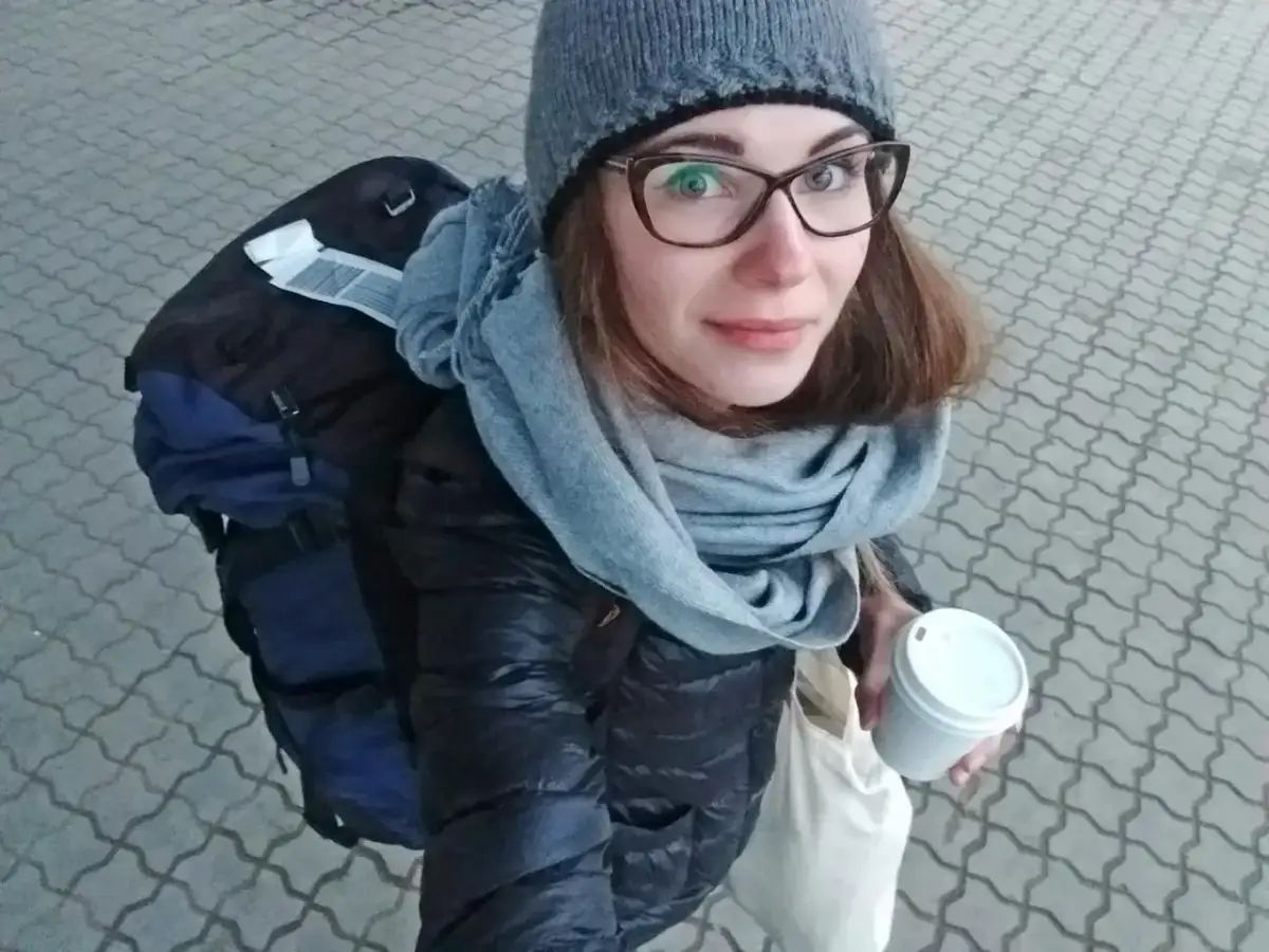 Kobieta w czapce i szaliku trzyma kubek kawy, obok plecak.