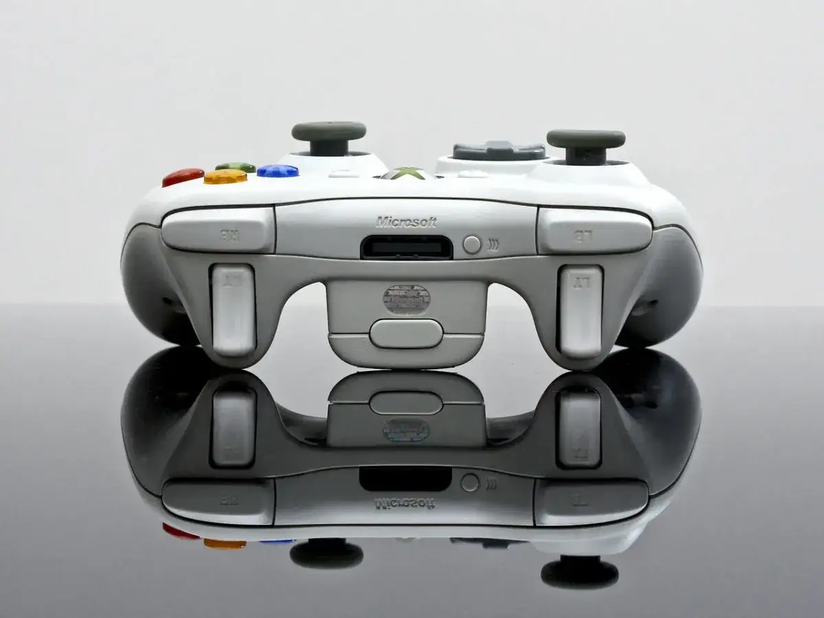 Biały kontroler Xbox 360 z kolorowymi przyciskami, odbijający się od czarnej powierzchni.