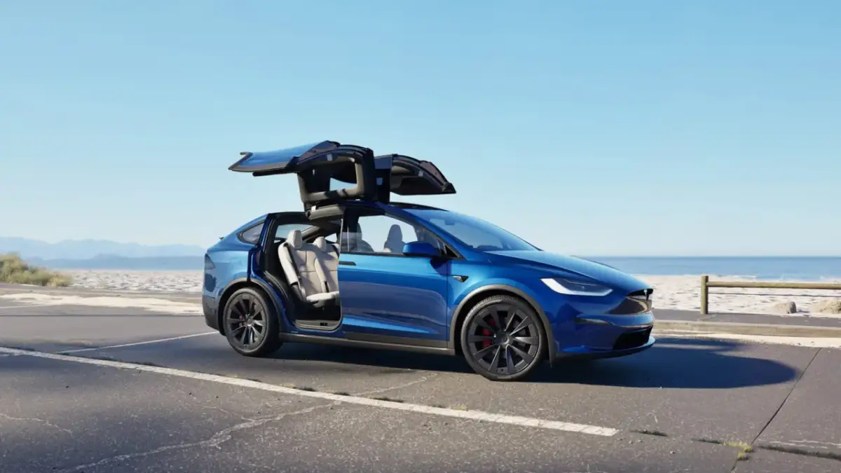 Niebieski Tesla Model X z otwartymi drzwiami typu falcon wing na tle plaży. Sprawdź aktualną tesla model x cena.