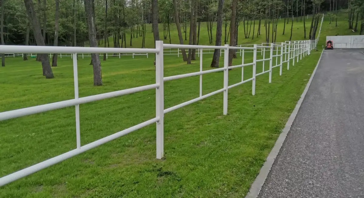 Biały, metalowy padok dla koni otacza zieloną łąkę z drzewami. W tle widać drogę i maszynę rolniczą.