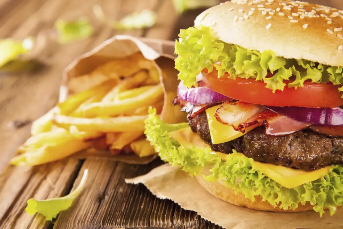 A close-up of a gourmet hamburger with lettuce, tomato, onion, bacon, and cheese, next to a bag of french fries.
