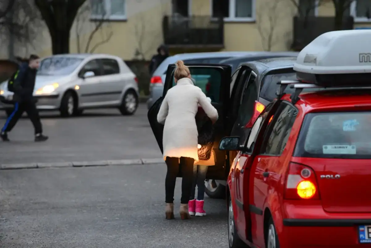 Kobieta z dzieckiem wsiada do samochodu, obok stoją inne auta.