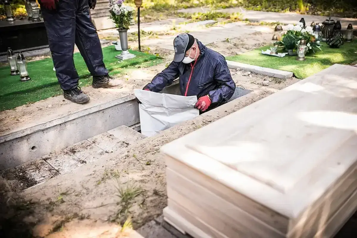 Pracownicy wykonują ekshumację, wyciągając biały worek z grobu. Obok leży drewniana trumna.