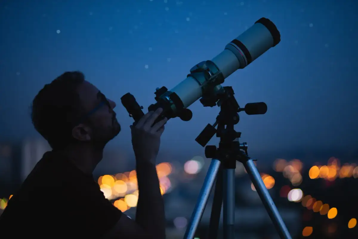 Mężczyzna obserwuje nocne niebo przez teleskop na tle rozświetlonego miasta.