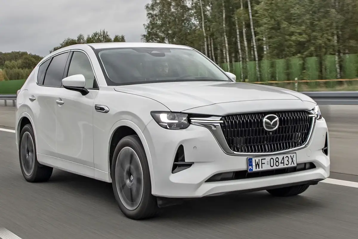 Biała Mazda CX-60 jedzie po drodze, otoczona drzewami. Sprawdź, jaka jest mazda cx 60 cena i wyposażenie.