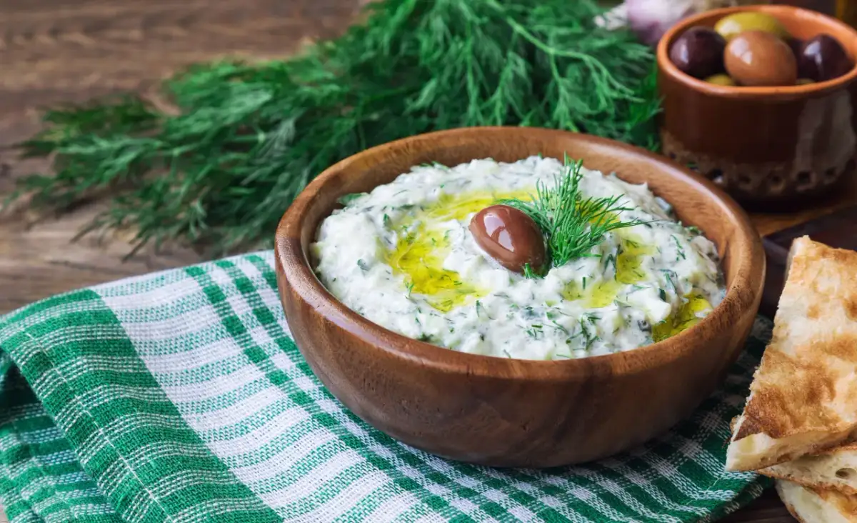 Oryginalny przepis na sos tzatziki: kremowy jogurt, świeży ogórek, czosnek, koperek i oliwa, ozdobiony oliwką.