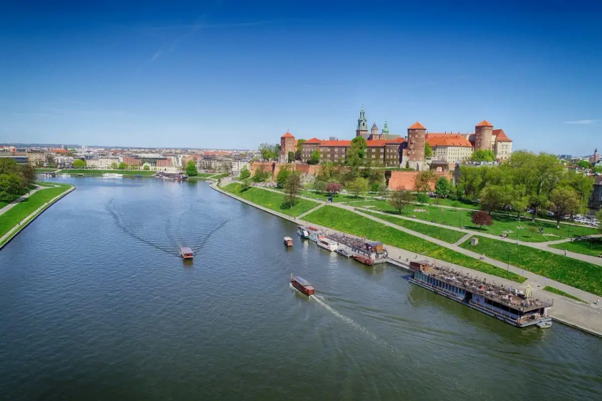 Widok na Wawel i rzekę Wisłę z łodziami. Słoneczny dzień zachęca do spacerów po zielonych bulwarach.