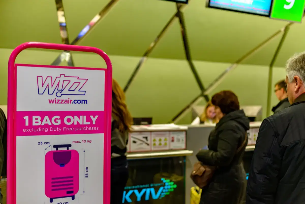 Tablica Wizz Air z informacją o limicie bagażu podręcznego i ludzie w tle.