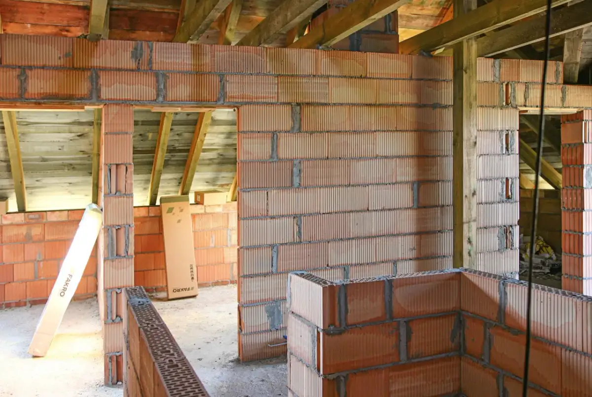 Budowa domu z bloczków z betonu komórkowego. Widać ściany z pustaków, drewniane belki stropowe i materiały budowlane, jak klej czy zaprawa do betonu komórkowego.