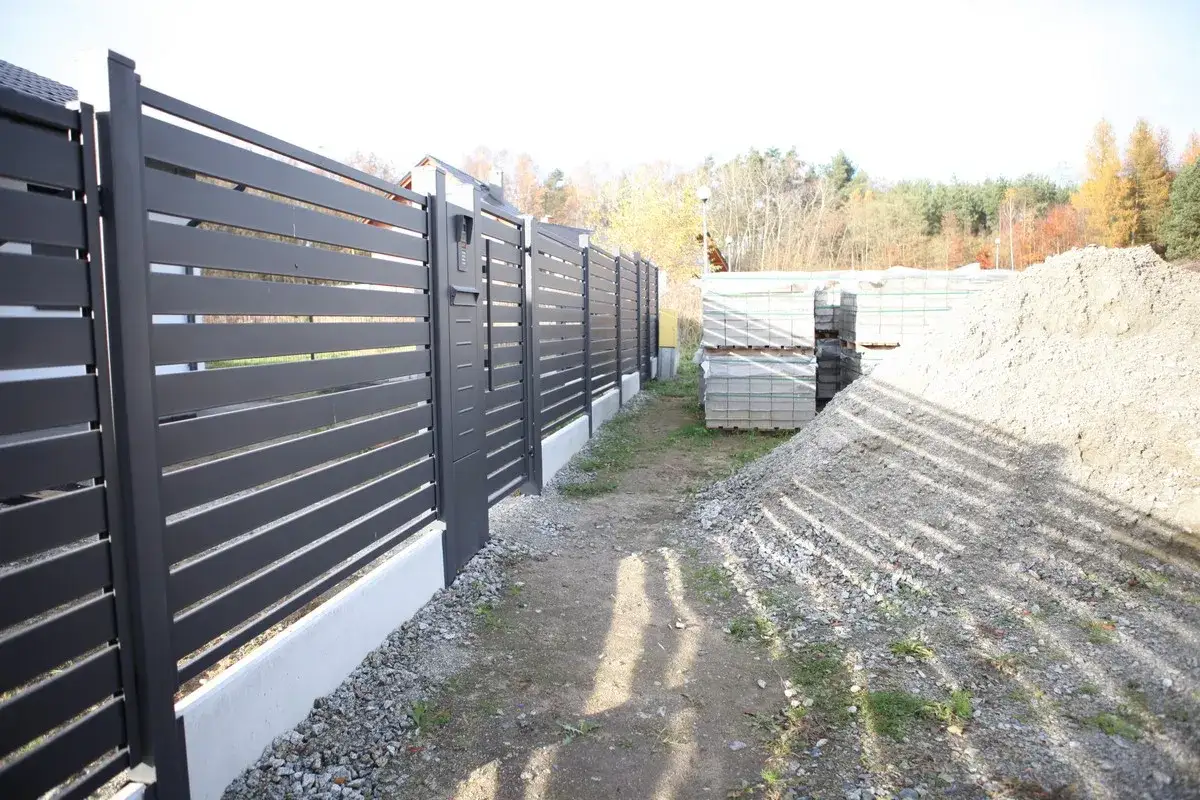 Schemat fundamentu pod bramę przesuwną. Widać nowoczesne ogrodzenie, materiały budowlane i hałdę żwiru.