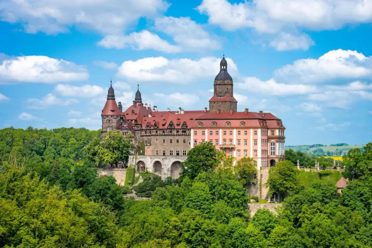 Imponujące pałace w Polsce, jak ten z czerwonym dachem i wieżami, otoczone bujną zielenią, przyciągają wzrok pod błękitnym niebem.