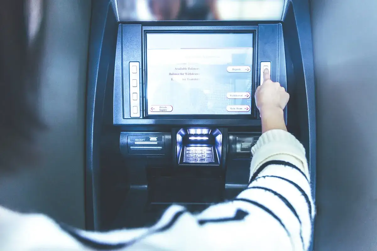 Ręka naciska przycisk na bankomacie. Ekran pokazuje opcje: "Deposit", "Withdrawal", "Main Menu". Czy z konta dziecka można robić przelewy?