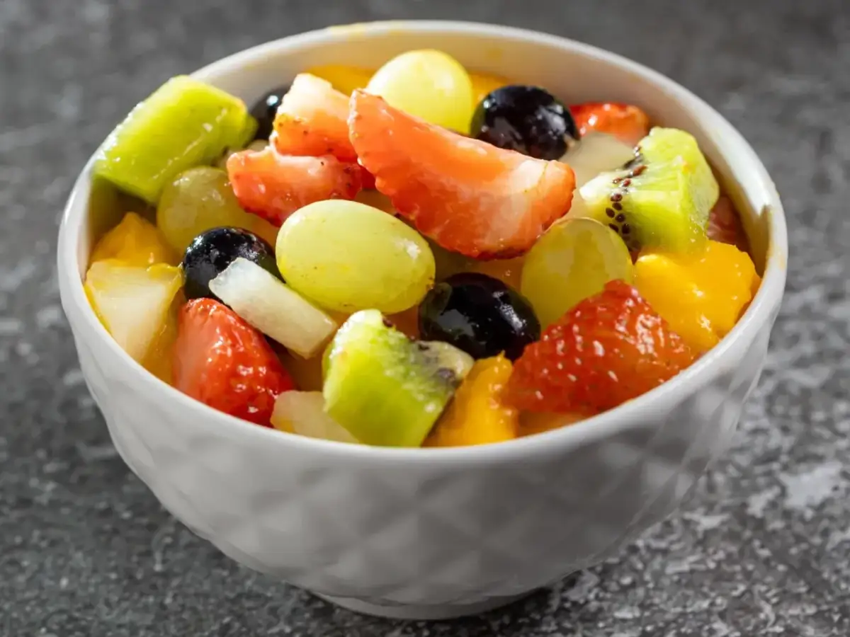 Kolorowa sałatka z owocami: truskawki, kiwi, winogrona, mango i borówki w białej miseczce.