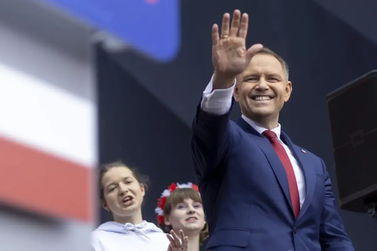 Uśmiechnięty polityk w garniturze pozdrawia tłum. W tle dwie młode kobiety, jedna z wiankiem na głowie. To może być część kampanii wyborczej, pozew w trybie wyborczym.