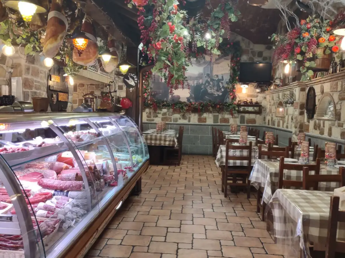 Wnętrze restauracji w stylu rustykalnym, z wiszącymi wędlinami i dekoracjami. W chłodni mnóstwo wyrobów, jak w najlepszym sklepie w Frascati.