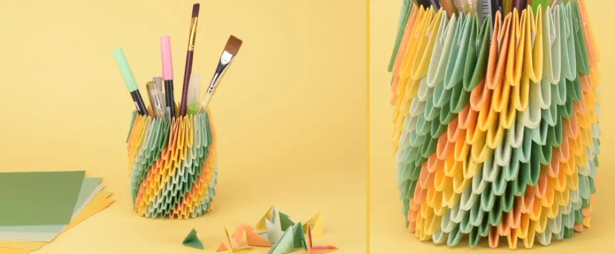 Bunter Papierkorb gefüllt mit Stiften und Pinseln, daneben Papierbögen und kleine Origami-Figuren.