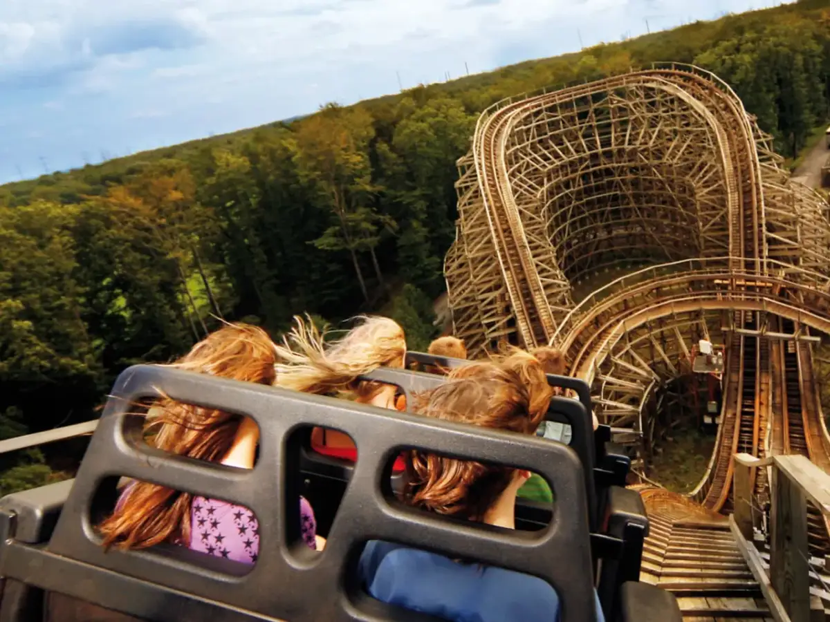 Fahrt mit einer Holzachterbahn, Menschen mit wehenden Haaren, im Hintergrund Wald und Himmel.