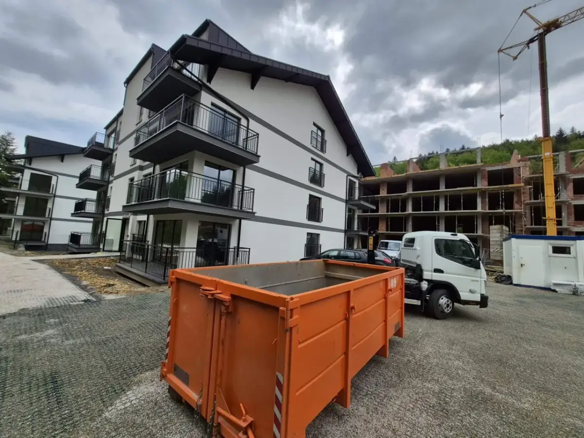 Pomarańczowy kontener na gruz przed nowym budynkiem i placem budowy.