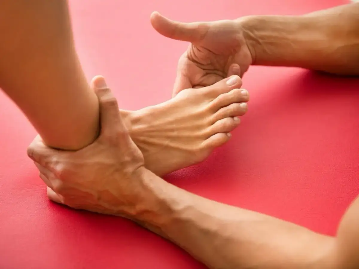 Dłonie masują stopę z widocznym haluksem na czerwonym tle.