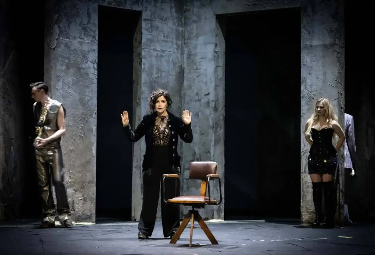 Na scenie, w ponurym otoczeniu, rozgrywa się wiśniowy sad spektakl. Kobieta w czerni wznosi ręce, obok stoi mężczyzna z saksofonem, a po drugiej stronie młoda kobieta w błyszczącej sukience.