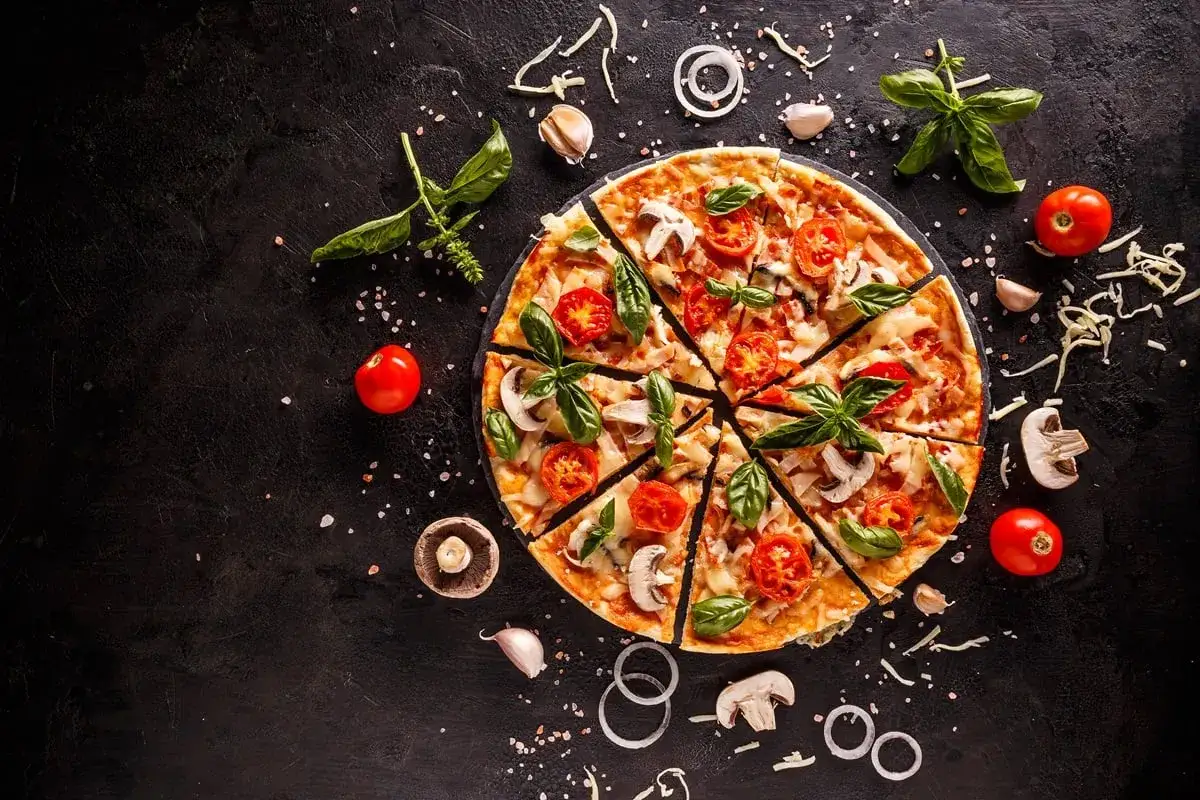 Pokrojona pizza z pomidorami, mozzarellą i bazylią na czarnym tle, otoczona składnikami.