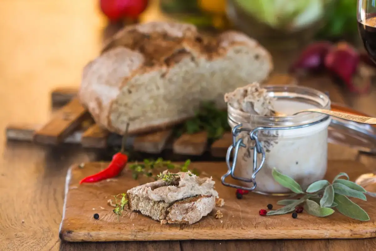 Pasztet na chlebie z ziołami, słoik pasztetu, chleb i papryczka chili na desce.