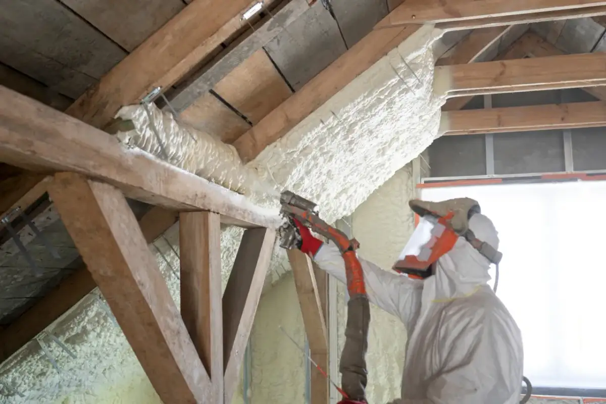 Izolacja poddasza pianą pur zamkniętokomórkową. Pracownik w kombinezonie ochronnym natryskuje pianę na drewniane belki konstrukcji dachu.