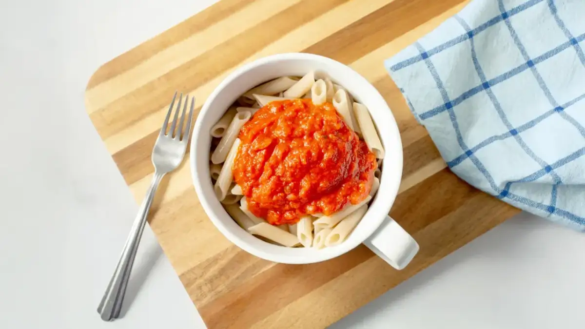 Makaron penne z sosem pomidorowym w białej misce, obok widelec i ściereczka.