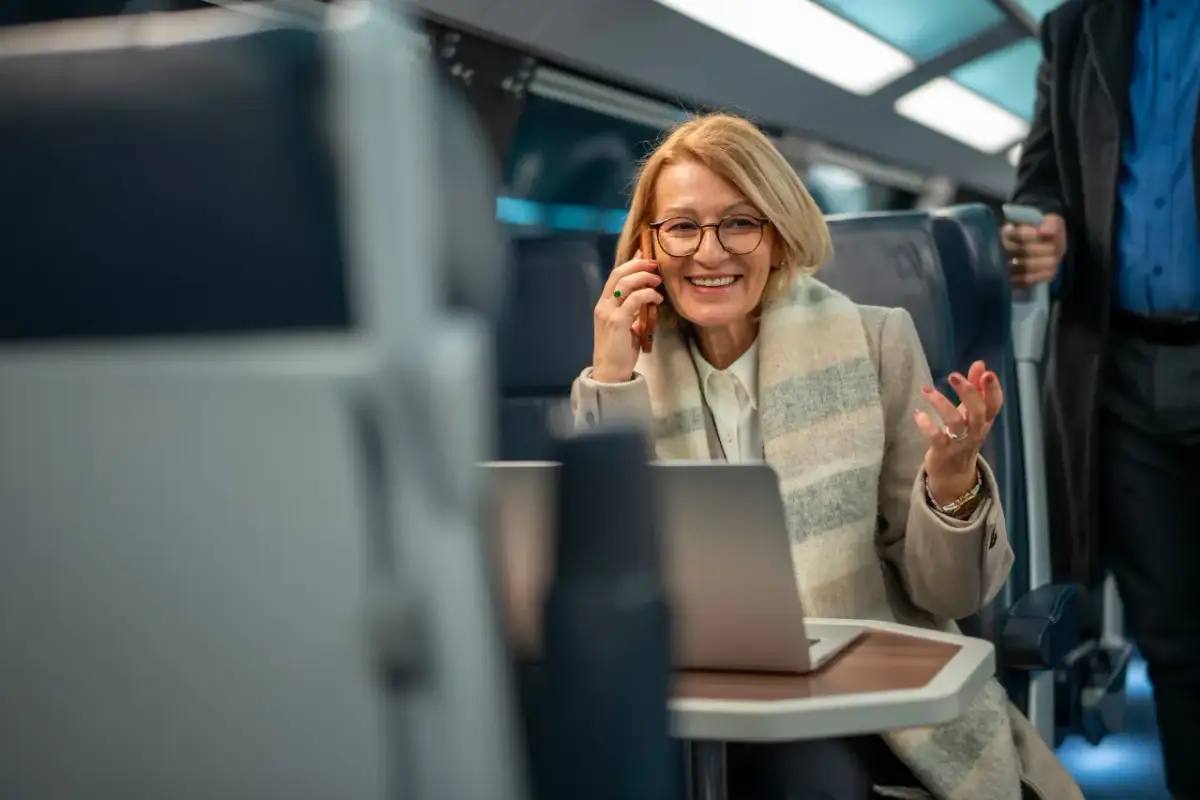 Kobieta w pociągu rozmawia przez telefon, pracuje na laptopie i gestykuluje.