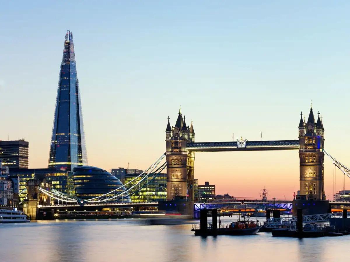 Widok na Tower Bridge i The Shard w Londynie o zmierzchu, z rzeką Tamizą i łodziami w tle.