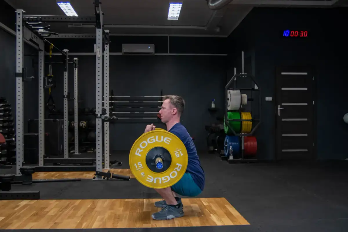 Mężczyzna wykonuje zercher squat z ciężarem w zgięciu łokci. Siłownia z wolnymi ciężarami i sprzętem do treningu siłowego.