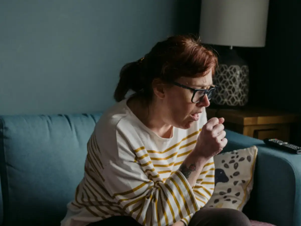 Kobieta w okularach siedzi na kanapie, kaszląc. Jej uporczywy suchy kaszel u dorosłego nie daje jej spokoju.