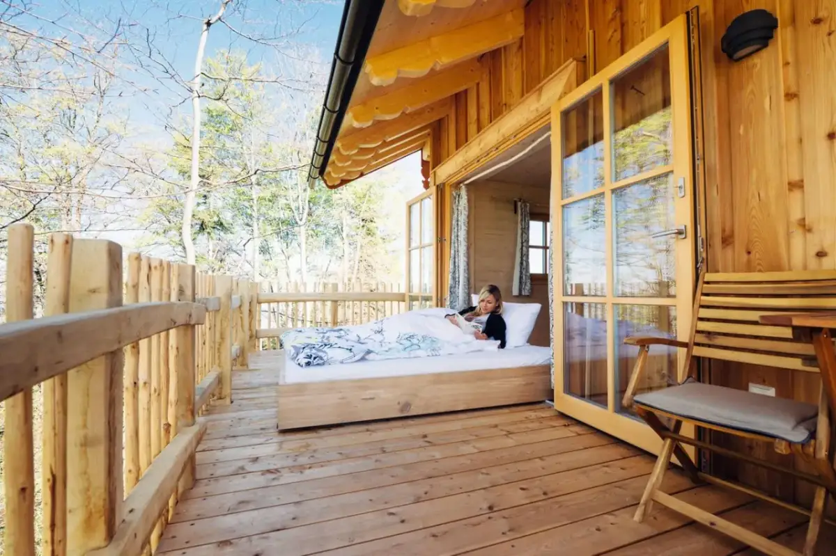Frau liest im Bett in einer Holzhütte mit offener Tür und Blick auf Bäume.
