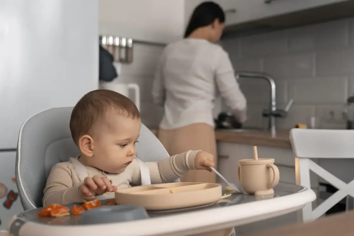 Dziecko siedzi w krzesełku do karmienia, próbując samodzielnie jeść. W tle mama przygotowuje posiłek.