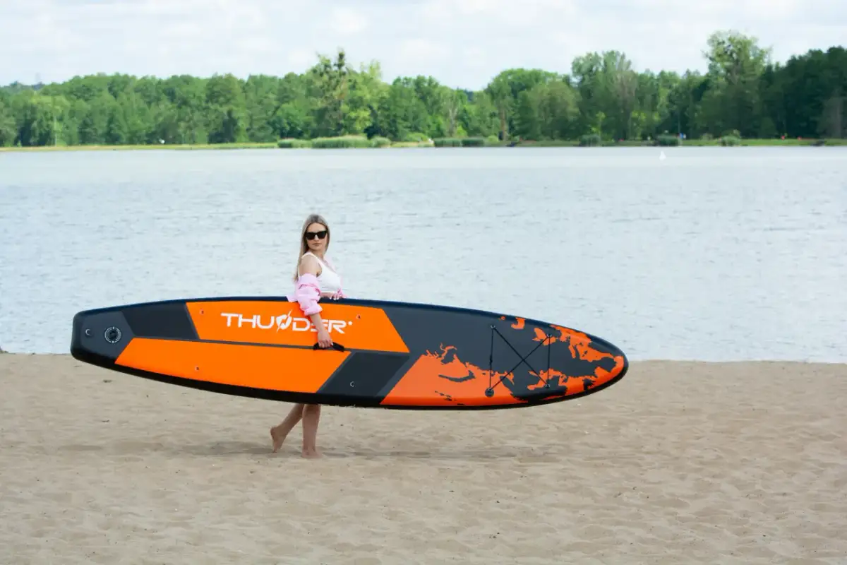 Kobieta z pomarańczowo-czarnymi deskami do pływania z wiosłem na piaszczystej plaży, gotowa do wodnych przygód.