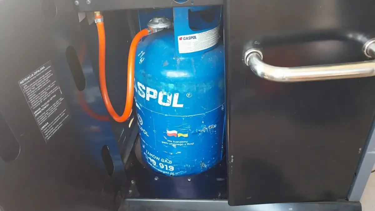 Niebieska butla z gazem marki GASPOL, podłączona wężem do grilla. To odpowiednia butla do grilla gazowego, zapewniająca zasilanie.