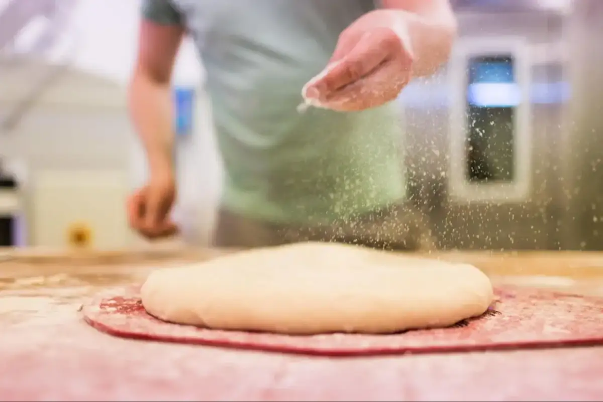 Ręka posypuje ciasto na pizzę mąką. Odpowiednia jaka mąka do pizzy to klucz do sukcesu.
