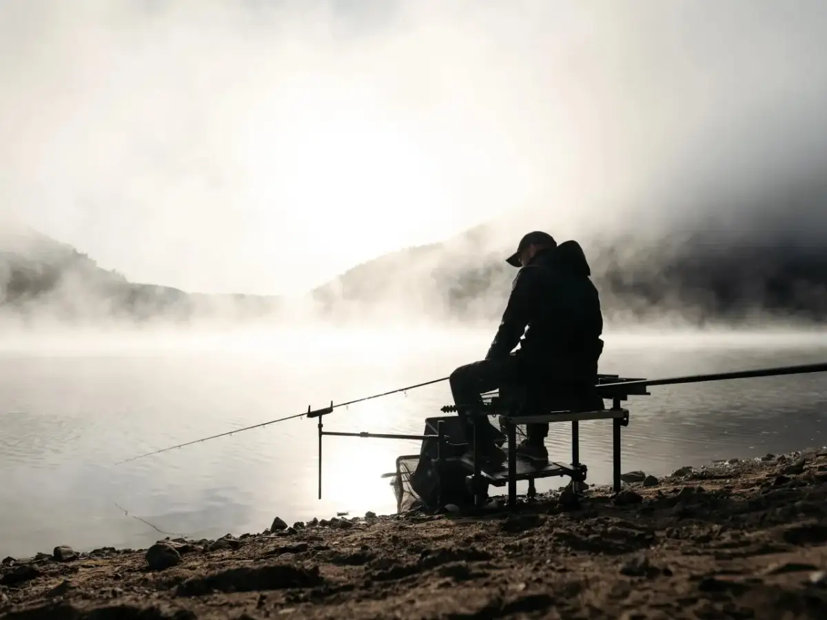 Wędkarz siedzi nad mglistym jeziorem o wschodzie słońca, łowiąc ryby.