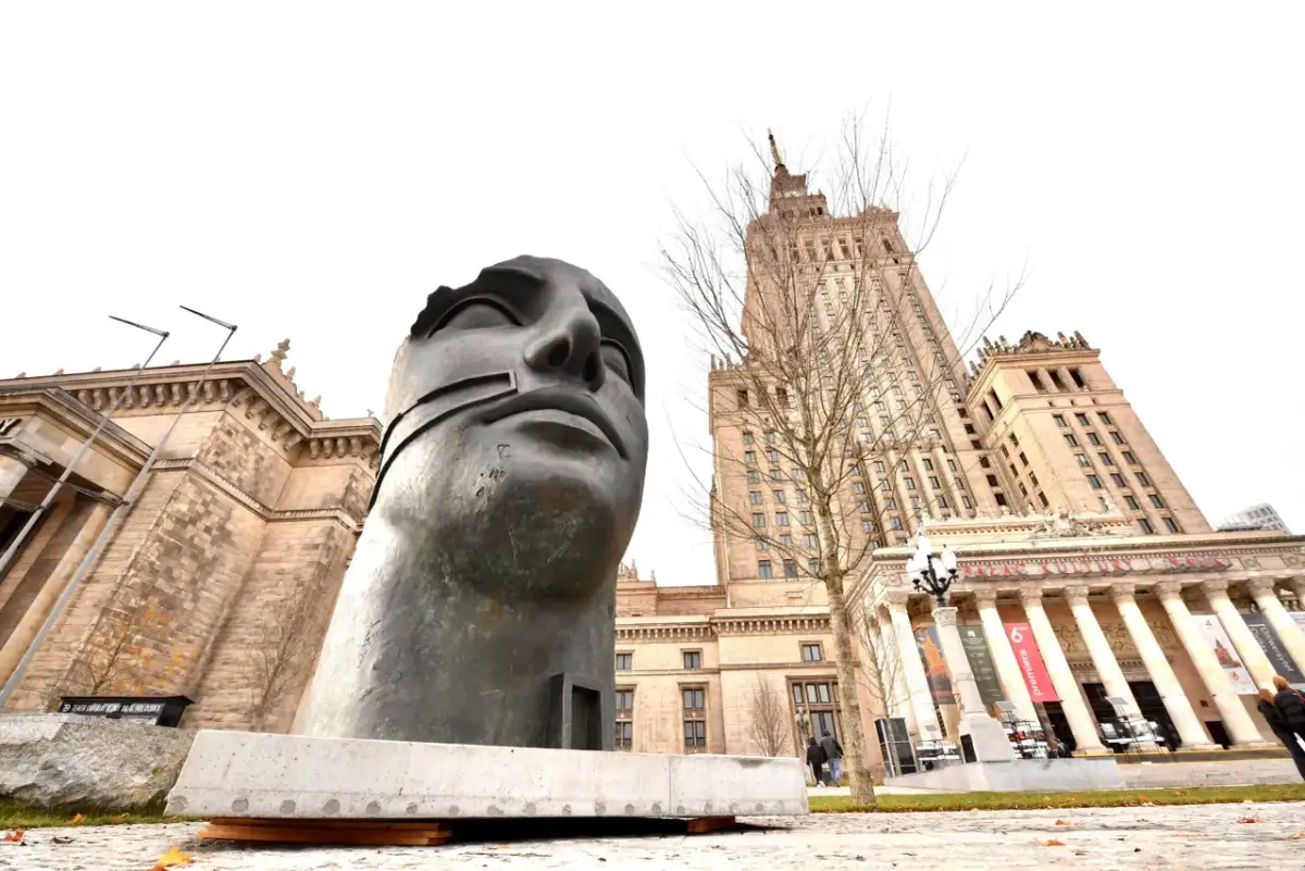 Monumentalna rzeźba głowy na tle Pałacu Kultury w Warszawie.
