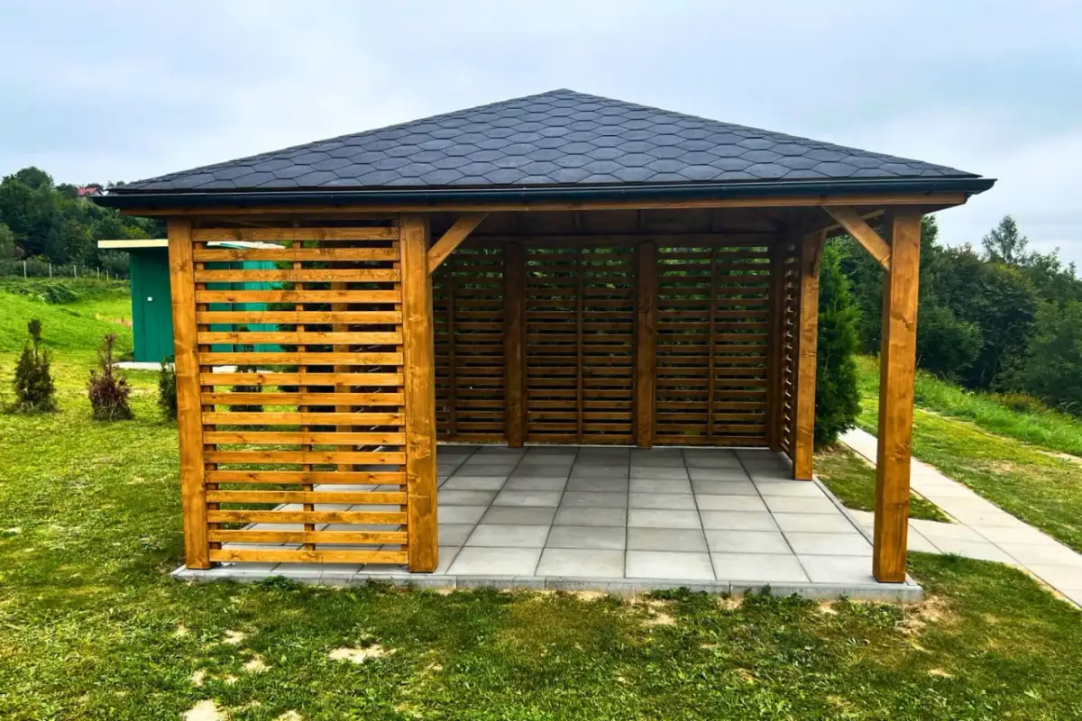 Drewniana altana z ażurowymi ściankami i ciemnym dachem. Idealne miejsce na relaks w ogrodzie, z pięknymi widokami.