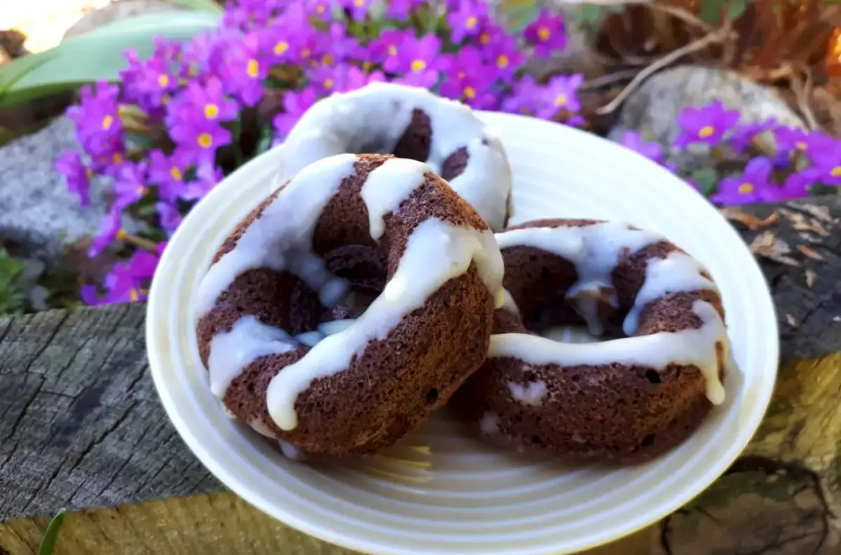 Trzy czekoladowe donuty brownie polane białym lukrem, na talerzu wśród fioletowych kwiatów.