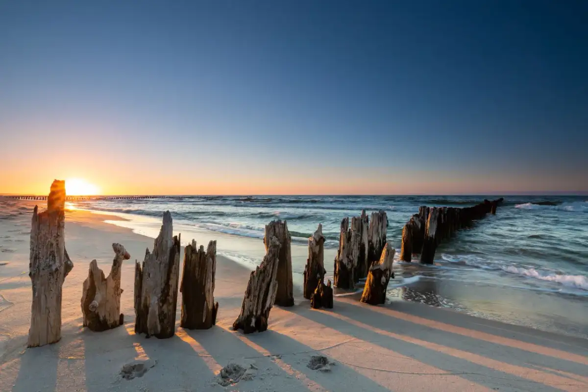 Zachód słońca nad Bałtykiem, z drewnianymi słupkami na plaży i falami.