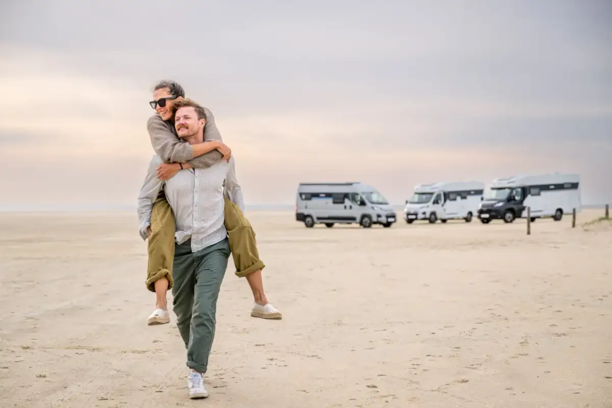 Szczęśliwa para na plaży, mężczyzna niesie kobietę na barana. W tle kilka kamperów, czyli vanów mieszkalnych, gotowych na przygodę.