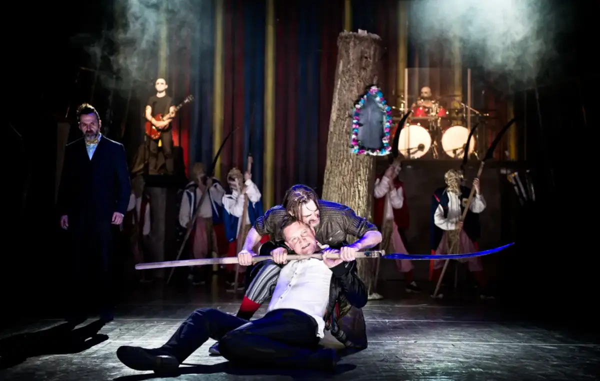 Jan Klata spektakle: dramatyczna scena z kosami i muzyką na żywo. Dwóch mężczyzn walczy na scenie, jeden klęczy, drugi go dusi.