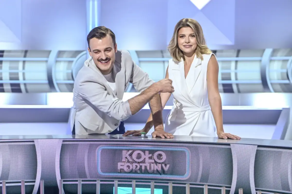 Uśmiechnięty mężczyzna i kobieta w białej sukience, którzy prowadzą Koło Fortuny, stoją przy pulpicie.
