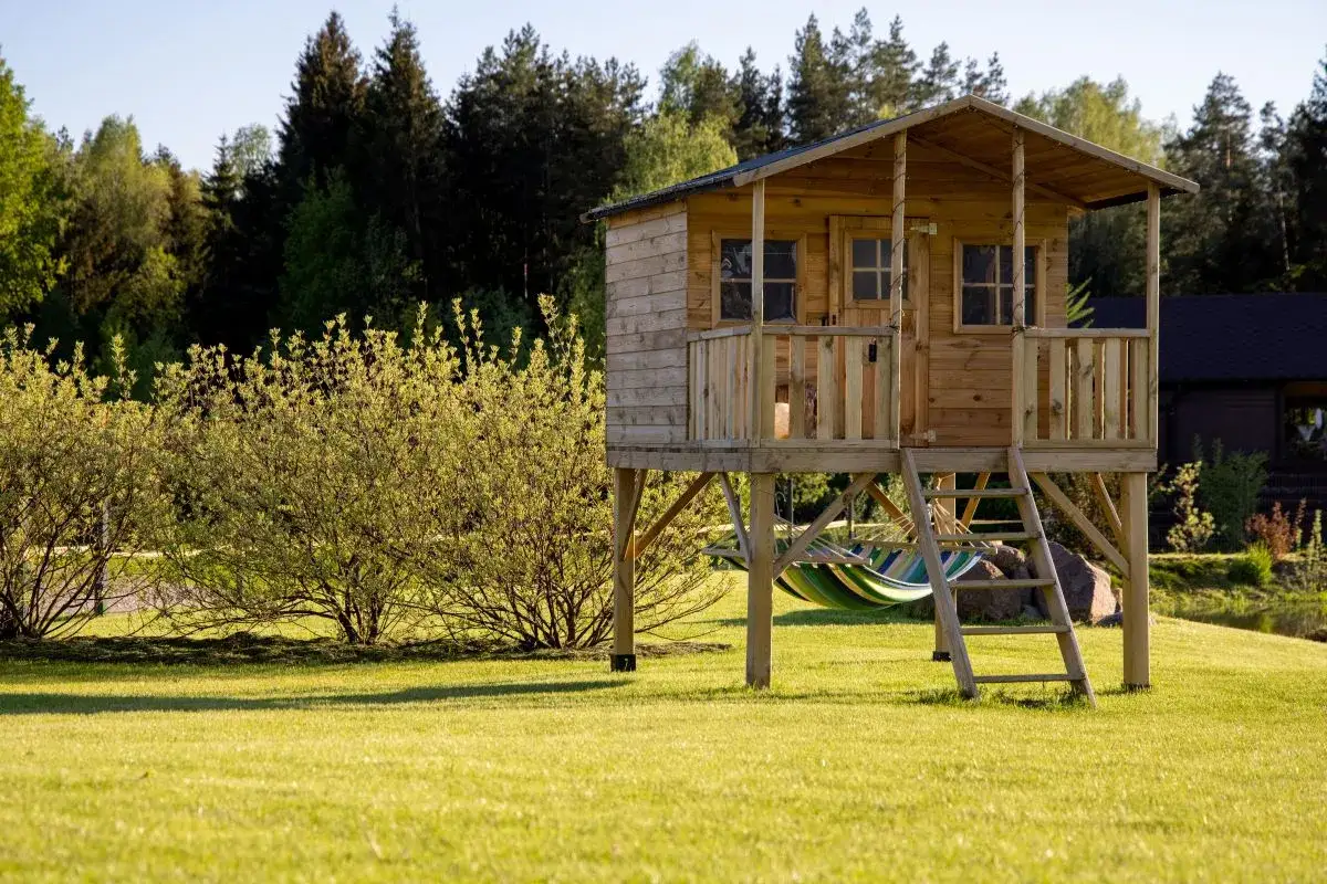 Drewniany domek na palach z drabinką i hamakiem na tle zieleni.