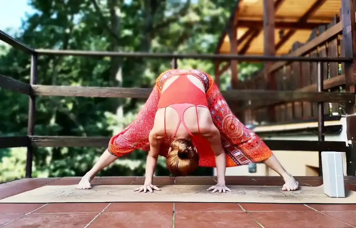 Kobieta w pomarańczowych spodniach i topie wykonuje skłon w jodze na macie na zewnątrz.