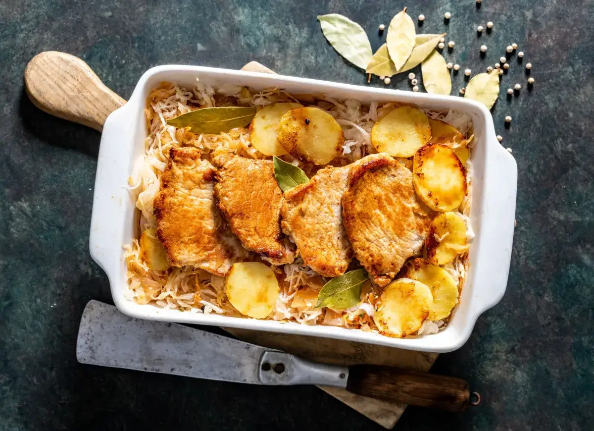 Pięć soczystych kotletów schabowych ułożonych na warstwie kiszonej kapusty, z plasterkami ziemniaków i liśćmi laurowymi. Pyszny schab w kapuście kiszonej.