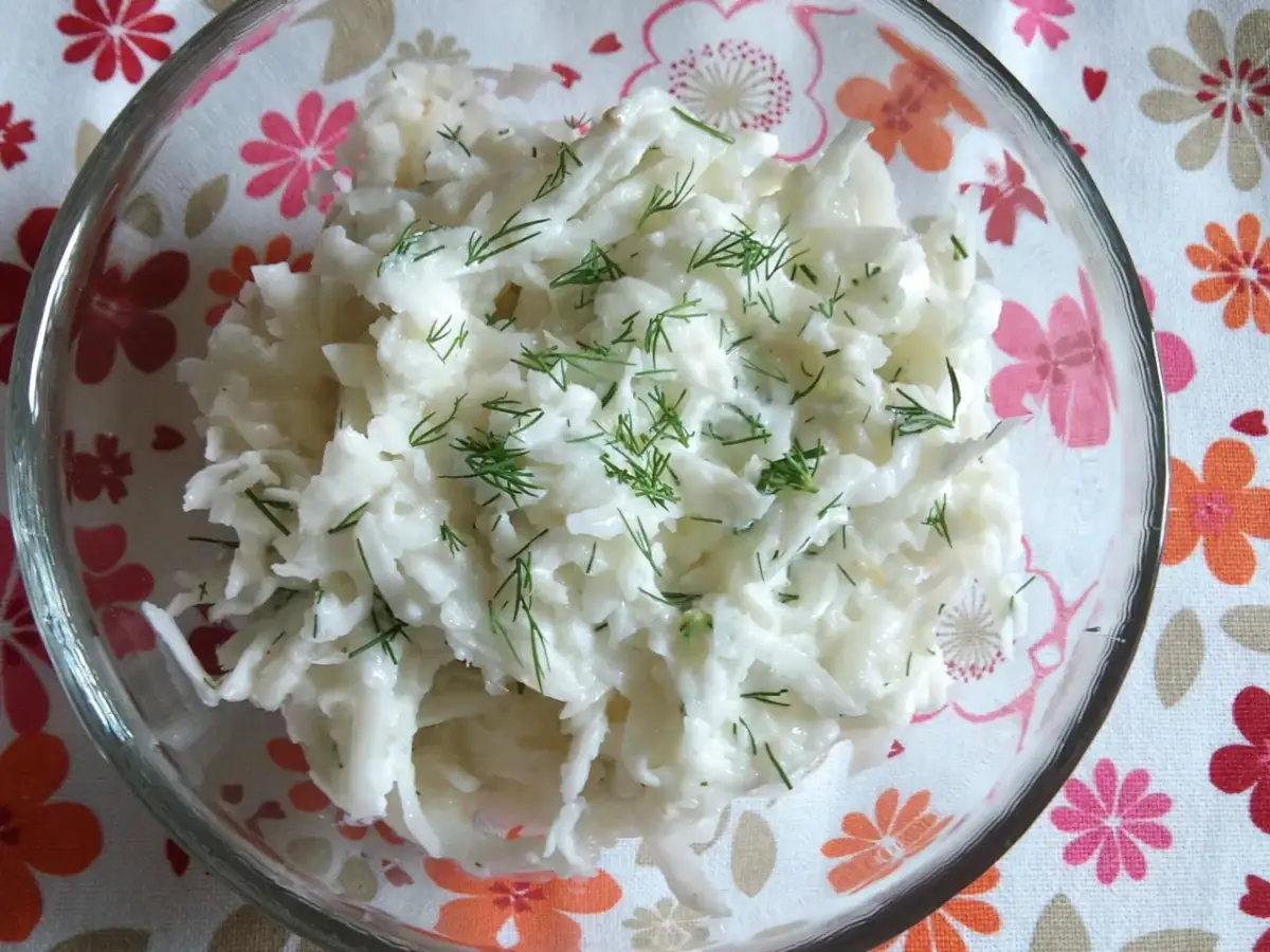 Pyszna surówka z białej rzodkwi, posypana świeżym koperkiem, w szklanej misce na kwiecistym obrusie.