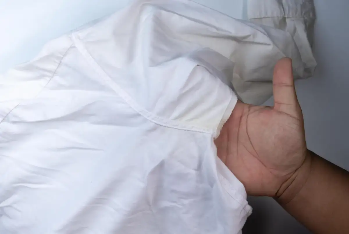 Dłoń pokazuje białą koszulę z żółtymi, twardymi plamami po antyperspirancie pod pachą.